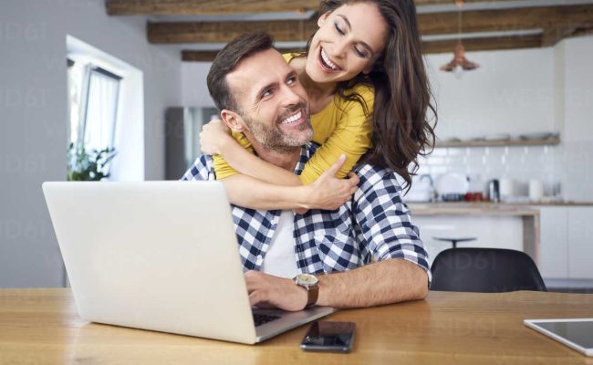 Happy couple sitting at dining table, embracing, using laptop